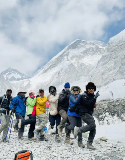 A group of jubilant trekkers posing at Everest Base Camp, elevation 5,364m, with towering snow-covered mountains and base camp tents behind them under a cloudy sky.