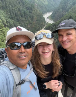 A selfie of two men and a woman, all wearing hats and sunglasses, smiling broadly on he route to Everest Base Camp trek with helicopter return with a deep, forested valley and a river visible behind them.