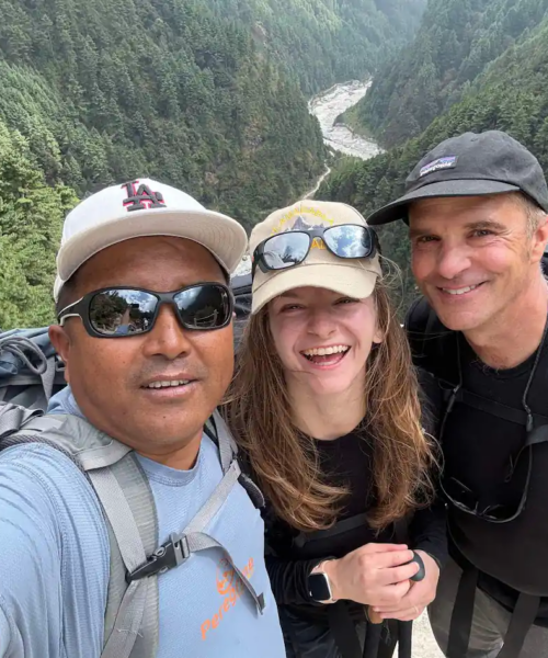 A selfie of two men and a woman, all wearing hats and sunglasses, smiling broadly on he route to Everest Base Camp trek with helicopter return with a deep, forested valley and a river visible behind them.