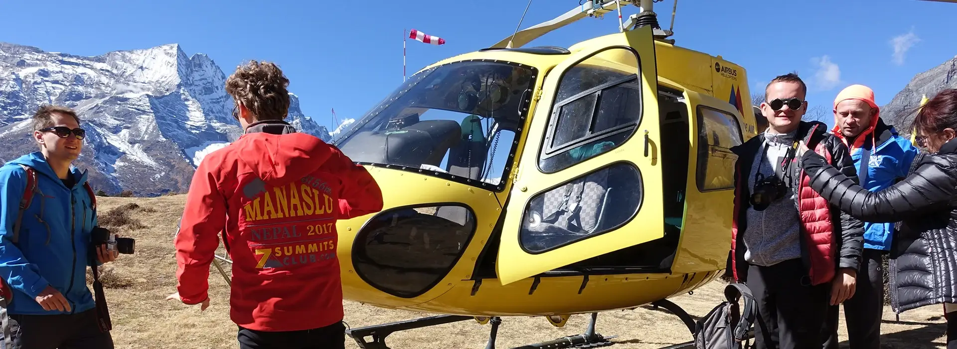 A bright yellow helicopter landed in a mountainous landscape, with several trekkers standing around it, some looking towards the helicopter and others towards the snow-capped peaks in the background.