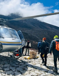 A group of trekkers approaches a silver helicopter parked on a rocky landing pad, with mountains rising in the background under a partly cloudy sky.