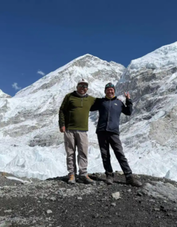 Two men posing happily on a rocky outcrop with vast snow-covered mountains and glaciers behind them under a clear blue sky.