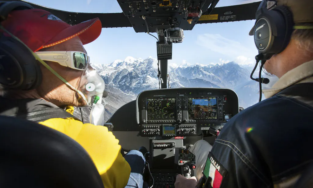 View from a helicopter cockpit showing a pilot and passenger, with the pilot wearing an oxygen mask, flying over snow-capped Himalayan mountains.
