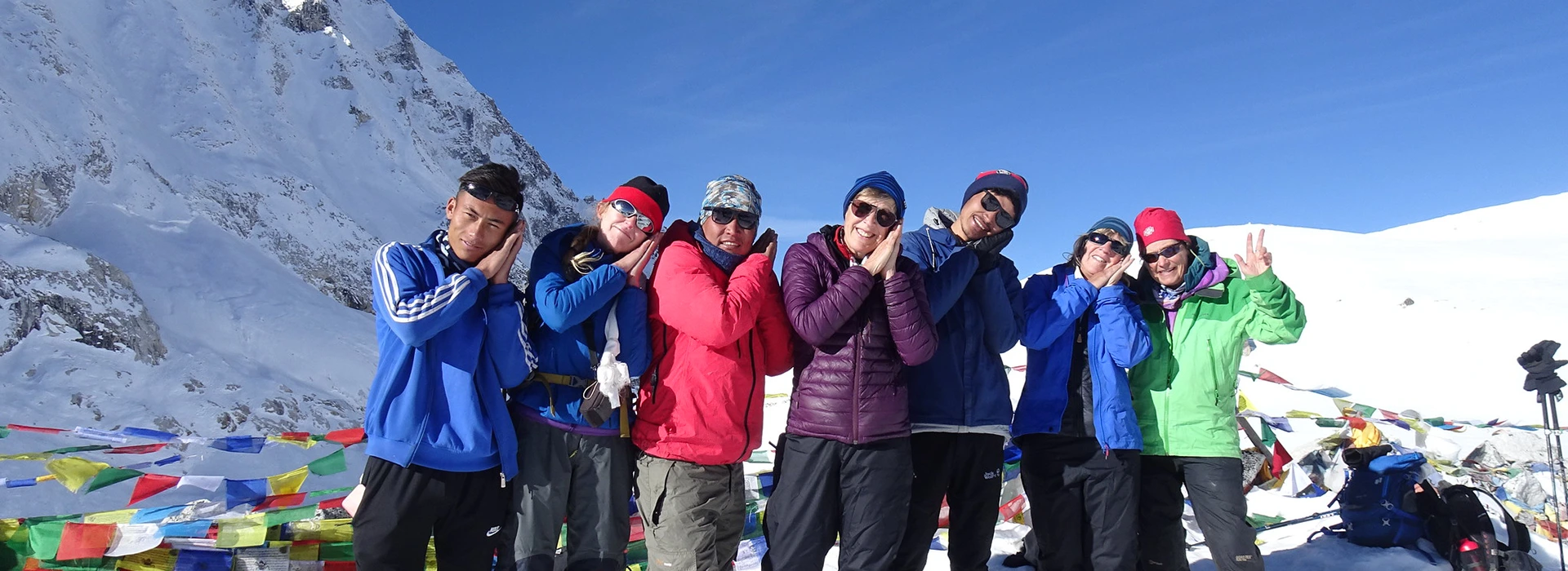 A large group of trekkers and their gear are spread across a vast, snow-covered mountain pass under a clear blue sky, with a cairn and prayer flags visible.