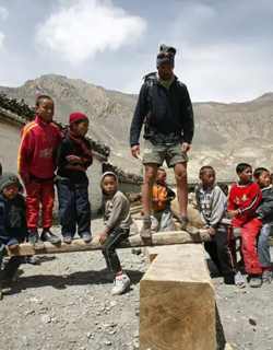 A tall trekker in hiking gear stands on a large wooden beam while surrounded by a group of local children in a rustic mountain village during an Annapurna Base Camp Hike.