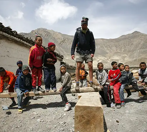 A tall trekker in hiking gear stands on a large wooden beam while surrounded by a group of local children in a rustic mountain village during an Annapurna Base Camp Hike.