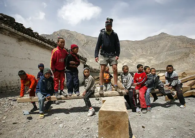 A tall trekker in hiking gear stands on a large wooden beam while surrounded by a group of local children in a rustic mountain village during an Annapurna Base Camp Hike.