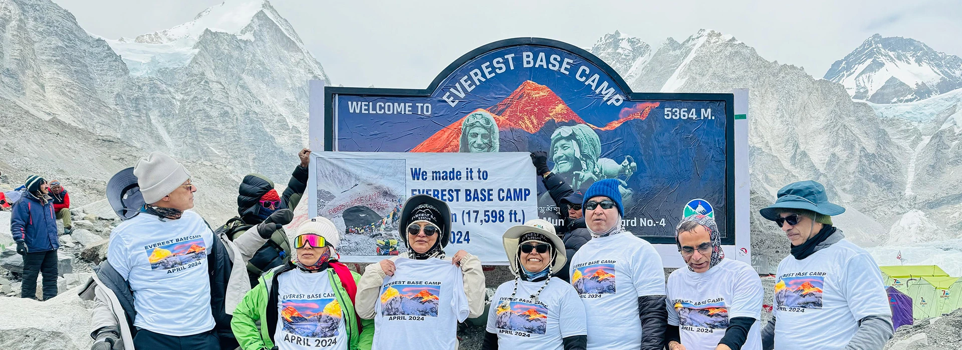 A group of jubilant trekkers in matching t-shirts stand in front of the Everest Base Camp sign, surrounded by snow-capped mountains and glaciers.