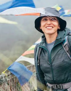 A smiling woman wearing a sun hat and hiking gear stands on a suspension bridge decorated with prayer flags during an Everest Base Camp Hike.