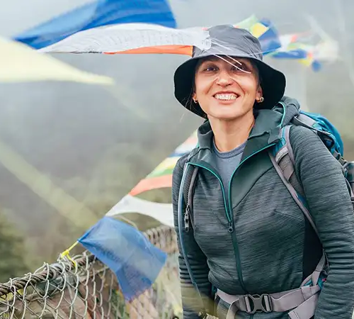 A smiling woman wearing a sun hat and hiking gear stands on a suspension bridge decorated with prayer flags during an Everest Base Camp Hike.