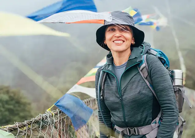 A smiling woman wearing a sun hat and hiking gear stands on a suspension bridge decorated with prayer flags during an Everest Base Camp Hike.