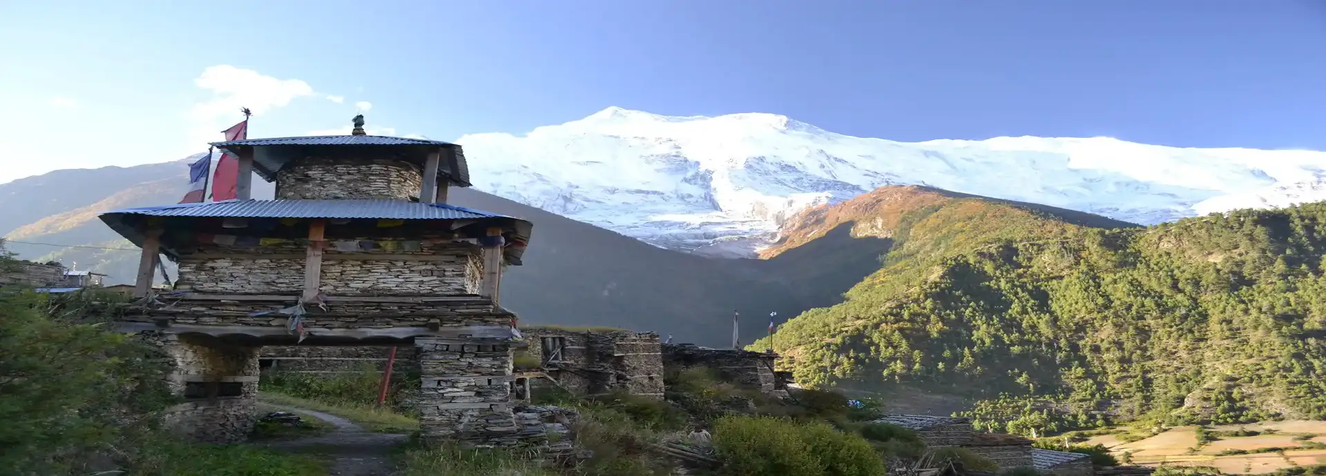 A multi-tiered stone gateway with a corrugated metal roof stands on a grassy path. In the background, a massive, snow-covered mountain dominates the horizon under a clear blue sky, flanked by sunlit green hills seen on the trail of Annapurna luxury lodge trek.
