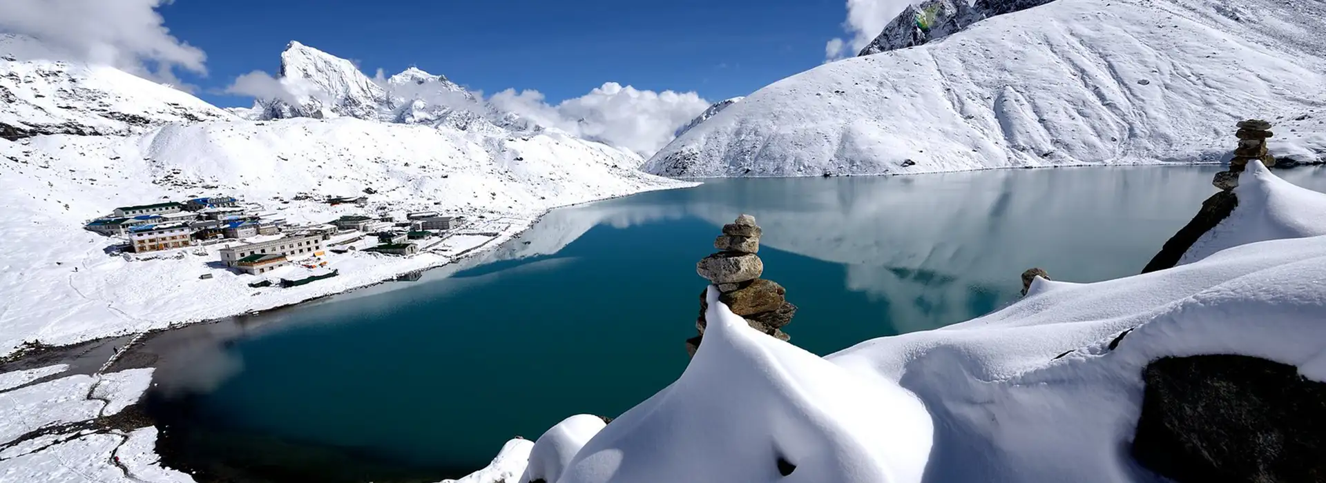 A stone cairn sits on a snowy ridge overlooking a turquoise glacial lake and a small village, captured during an Everest Three Passes Trek.