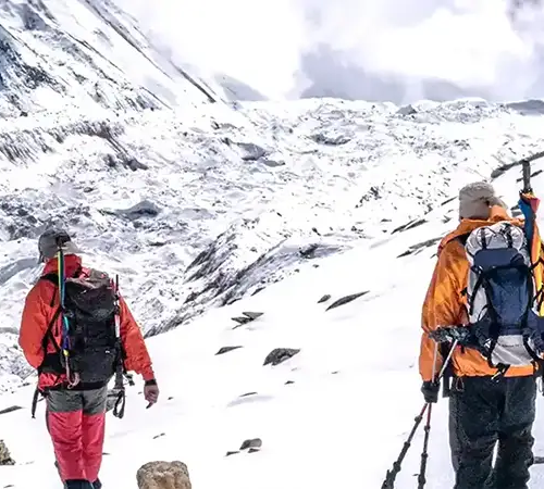 Two mountaineers in orange and red jackets carry heavy backpacks while Lobuche Peak climbing across a snowy, rocky mountain pass.