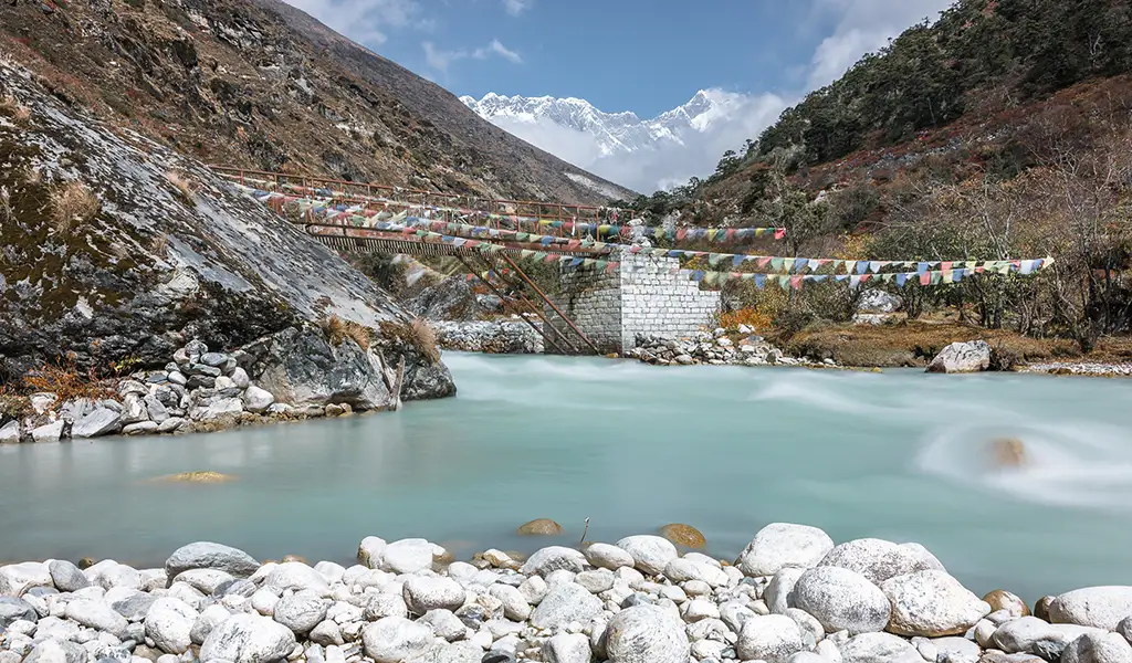 A view of a turquoise river and a suspension bridge decorated with prayer flags near Pangboche during an Everest Base Camp Trek for beginners.