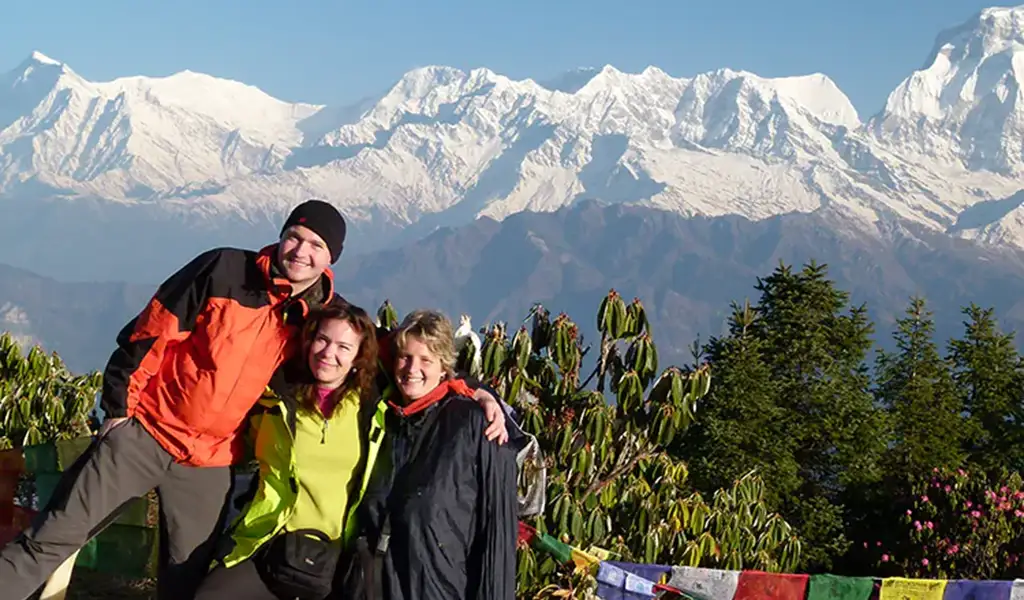 Three smiling trekkers pose together in front of the snow-capped Annapurna and Dhaulagiri mountain ranges at the Poon Hill viewpoint in Nepal.