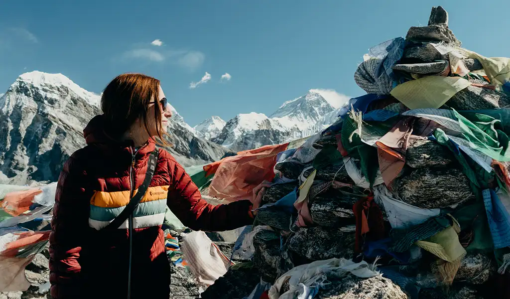 A hiker stands among Buddhist prayer flags at the summit of a rocky mountain pass, looking out over a turquoise lake and snowy peaks during an Everest Three Passes Trek.