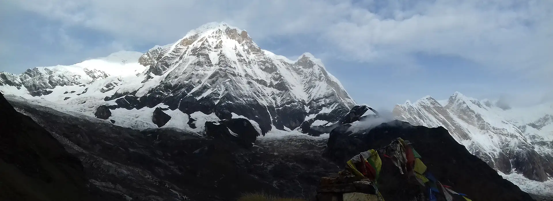 A wide mountain panorama showing massive, jagged peaks covered in snow and ice. Dark, rocky foothills lead up to the white summits under a cloudy sky, with colorful prayer flags visible on a small stone mound in the lower right foreground.