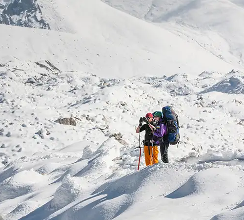 Two hikers with large backpacks and ice axes stand on a vast, undulating field of white snow and ice during a Gokyo Trek in the Himalayas.