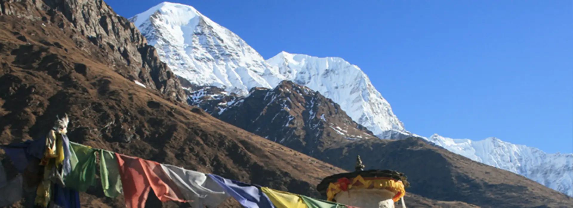 Bright Buddhist prayer flags stretch across the foreground of a mountain landscape. In the background, a white stupa stands before massive, snow-covered Himalayan peaks under a clear blue sky.