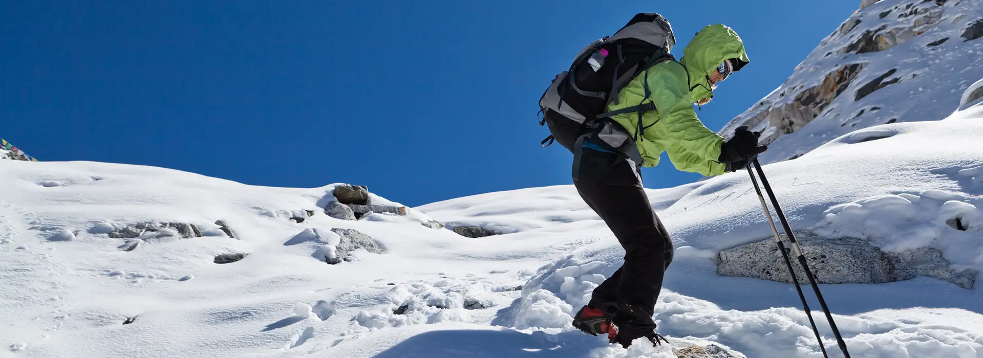 A trekker in a green jacket and backpack uses trekking poles to navigate a snowy slope during an Everest Base Camp Trek for beginners.