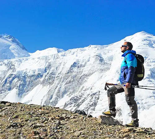A hiker wearing a bright green down jacket and a grey backpack uses trekking poles to climb a steep snow covered slope during an Everest Base Camp Trek for beginners.