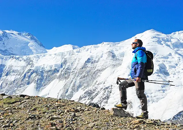 A hiker wearing a bright green down jacket and a grey backpack uses trekking poles to climb a steep snow covered slope during an Everest Base Camp Trek for beginners.