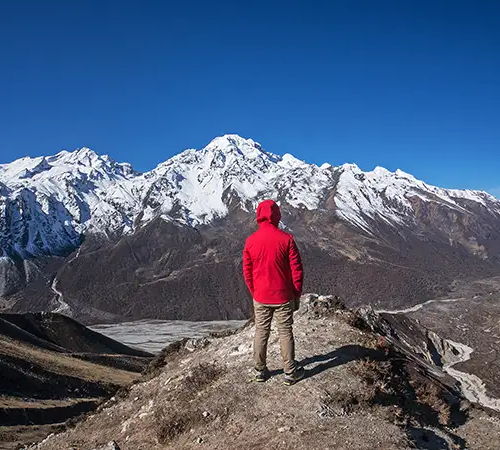 A trekker in a red jacket and brown pants stands on a rocky outcrop, looking out over the Langtang Valley toward snow-capped Himalayan peaks under a clear blue sky.