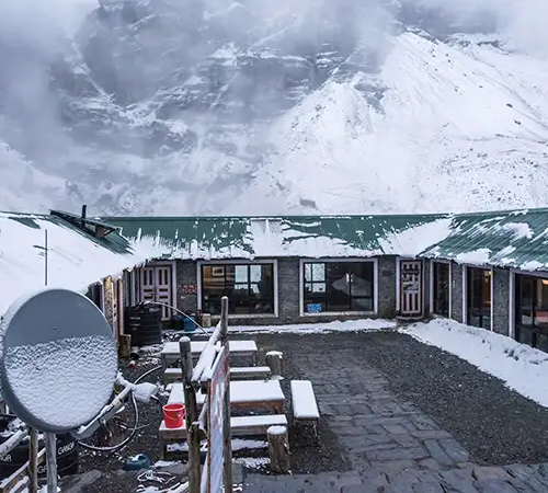 A stone trekking lodge with a green roof forms a U-shaped courtyard covered in fresh snow. In the background, massive, jagged rock faces and snow-dusted slopes disappear into low-hanging clouds and mist.