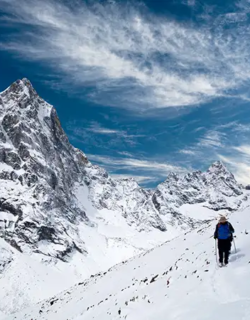 A lone hiker with trekking poles walks across a vast, snow-covered mountain slope during an Everest Panorama Trek, with a sharp, rocky peak and a wispy blue sky in the background.