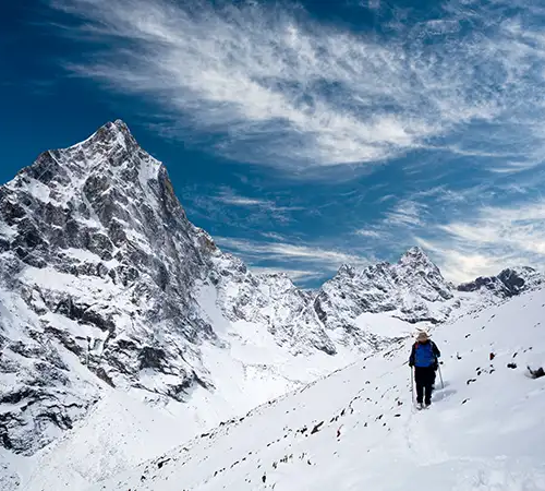 A lone hiker with trekking poles walks across a vast, snow-covered mountain slope during an Everest Panorama Trek, with a sharp, rocky peak and a wispy blue sky in the background.