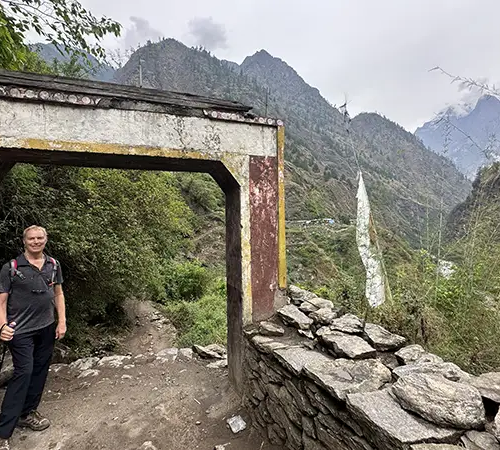A man with a hiking pole stands under a weathered stone archway on a mountain trail. In the background, steep green hills and the snow-capped peaks of the Ganesh Himal range rise under a cloudy sky during the Ganesh Himal Trek.