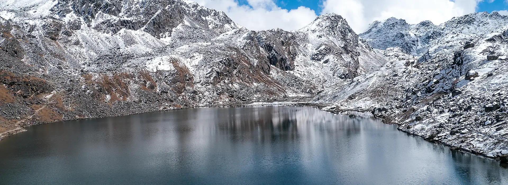 A serene view of Gosaikunda Lake along the Langtang Gosaikunda Trek in Nepal, showing the alpine lake reflecting snow-capped mountains under a blue, cloudy sky.