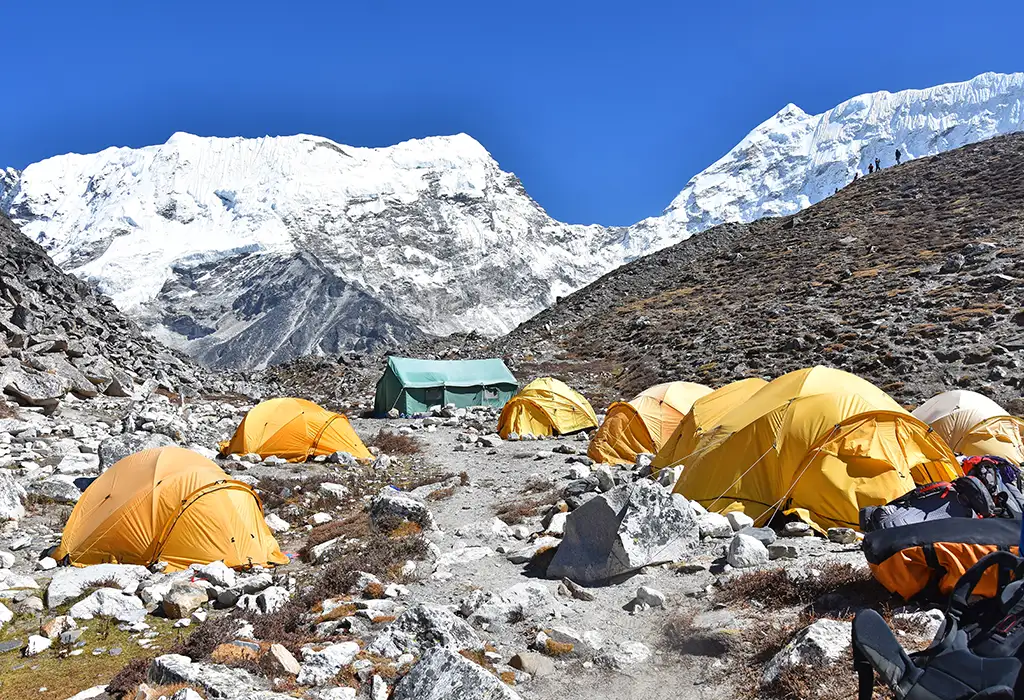 A rocky mountain base camp filled with yellow and green tents sits in a valley, serving as a staging point for Island Peak climbing under a clear blue sky.