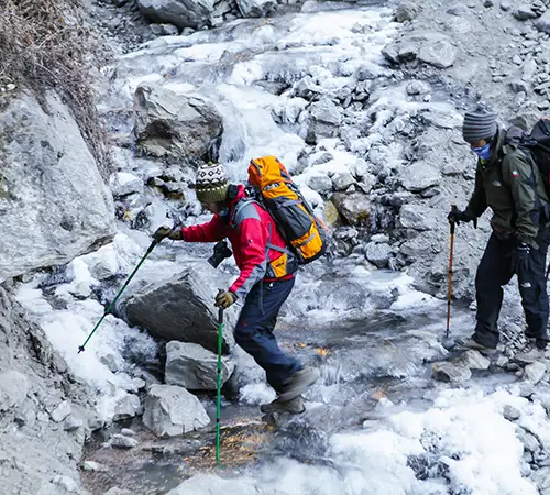 Three hikers with large backpacks and trekking poles carefully cross a rocky, partially frozen stream in a rugged mountain landscape.
