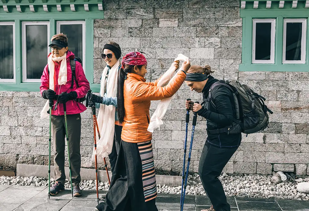 A local woman in an orange jacket and traditional striped apron places a white khata scarf around the neck of a hiker