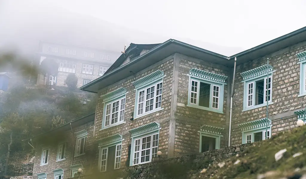 A traditional Himalayan stone lodge with decorative teal window frames sits on a misty hillside, a common resting point for those preparing for Island Peak climbing.