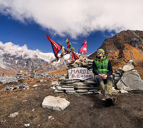 A person in a green puffer vest sits on a stone structure next to a sign that reads "MARDI B.C. 4500." Two large Nepalese flags and several prayer flags flutter from a wooden pole behind them, with a dramatic backdrop of autumn-colored hills and snowy peaks in Mardi Himal Trek.