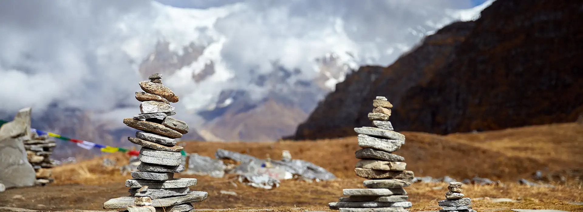 Several stacks of flat grey stones, or cairns, stand in a brown grassy field. In the blurry background, a massive snow-covered mountain peak is partially hidden by thick white clouds.
