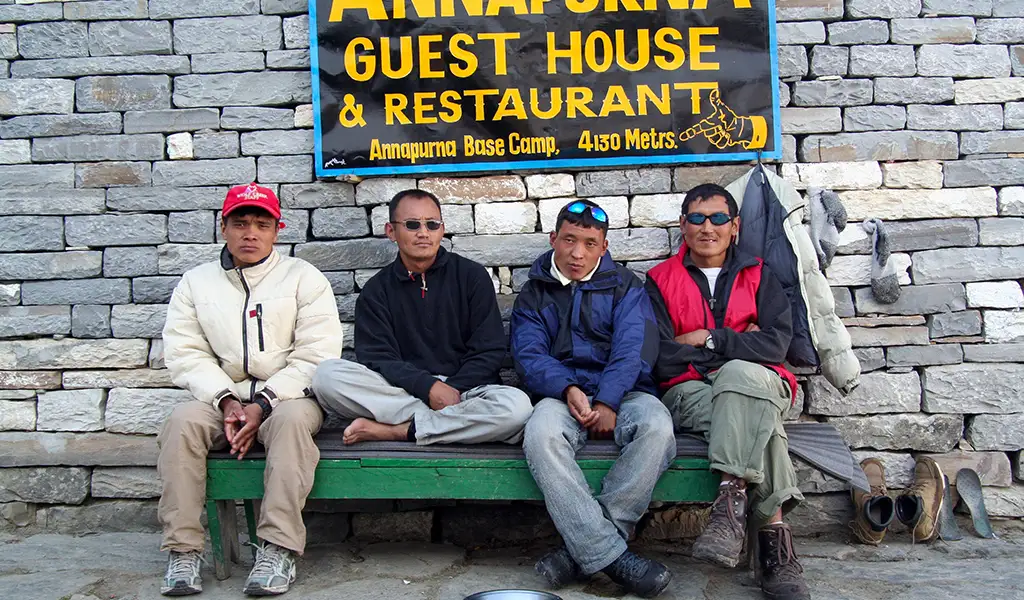 Four men sit on a green bench outside a stone building beneath a sign reading “Annapurna Guest House & Restaurant” at Annapurna Base Camp.