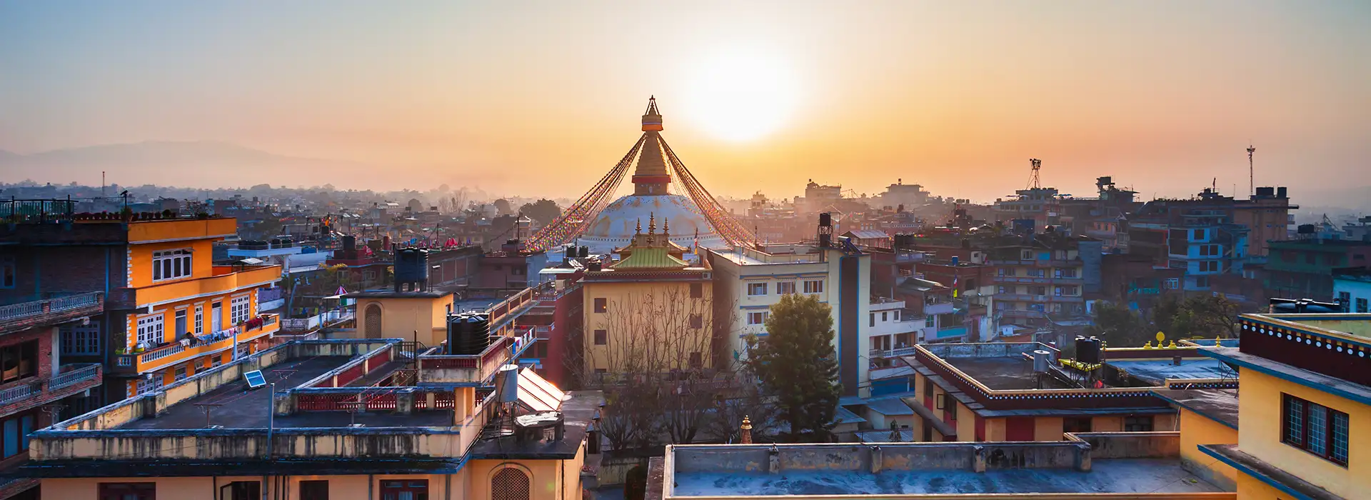 A wide panoramic shot of the golden Swayambhunath Stupa rising above the Kathmandu city skyline during a hazy sunset.