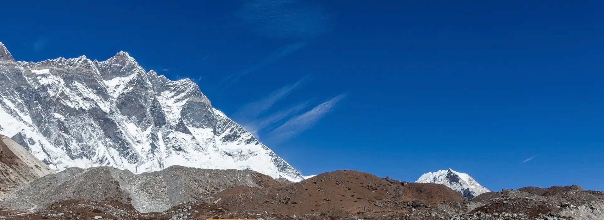 A wide landscape featuring a massive, triangular mountain peak covered in snow and ice on the left. In the foreground, brown rocky hills lead toward the mountain, while a smaller snowy peak is visible on the far right under a deep blue sky with light wispy clouds.