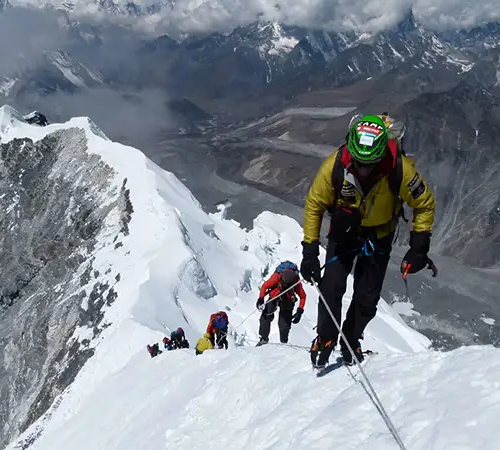 A line of climbers in bright technical gear follows a fixed rope up to the three peaks climbing route. Below them, a vast glacial valley stretches toward distant, cloud-shrouded peaks under a grey sky.