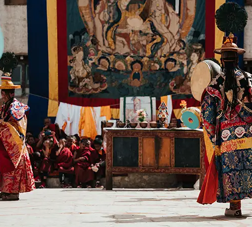 Two dancers in ornate, colorful silk robes and wide-brimmed hats perform with drums in a courtyard. A large thangka painting hangs in the background above an altar, while a crowd of monks in maroon robes watches the ceremony during upper mustang trek.