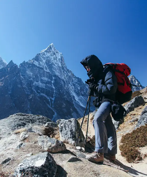 A hiker in a navy down jacket and red backpack uses trekking poles on a rocky trail with a sharp, snowy peak behind them during a Gokyo Lake Everest Base Camp Trek.
