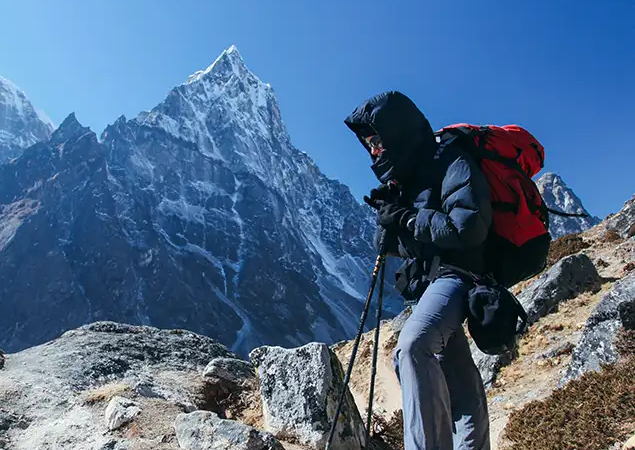 A hiker in a navy down jacket and red backpack uses trekking poles on a rocky trail with a sharp, snowy peak behind them during a Gokyo Lake Everest Base Camp Trek.