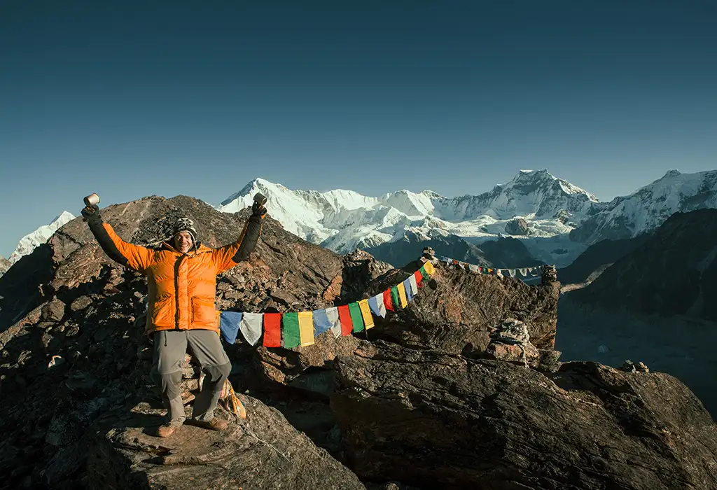 A triumphant trekker in an orange puffer jacket raises two mugs at the rocky summit of Gokyo Ri, marking a milestone of their Gokyo Trek with prayer flags and the Himalayas in the background.