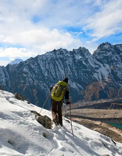 A trekker with a green backpack and trekking poles walks down a steep, snow-covered mountain slope during Manaslu Circuit Trek.
