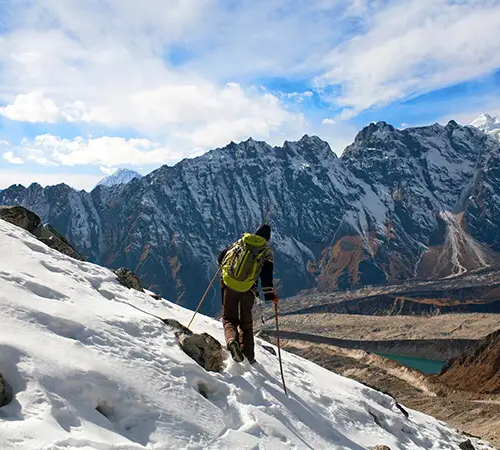 A trekker with a green backpack and trekking poles walks down a steep, snow-covered mountain slope during Manaslu Circuit Trek.