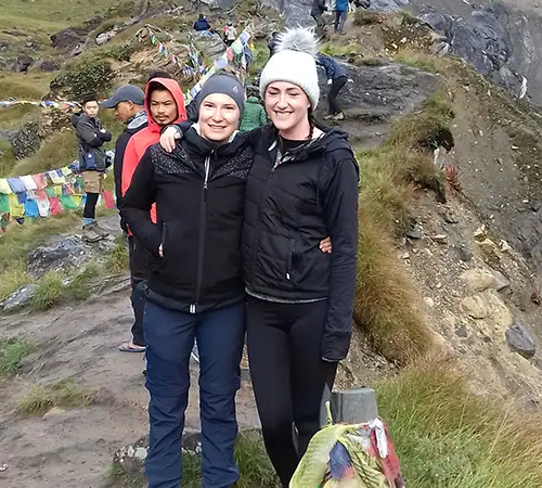 Two women in hiking jackets and beanies stand with their arms around each other on a narrow dirt path. Colorful prayer flags line the trail behind them, and other hikers are visible in the background against a steep mountain slope in the short Annapurna Base Camp Trek.